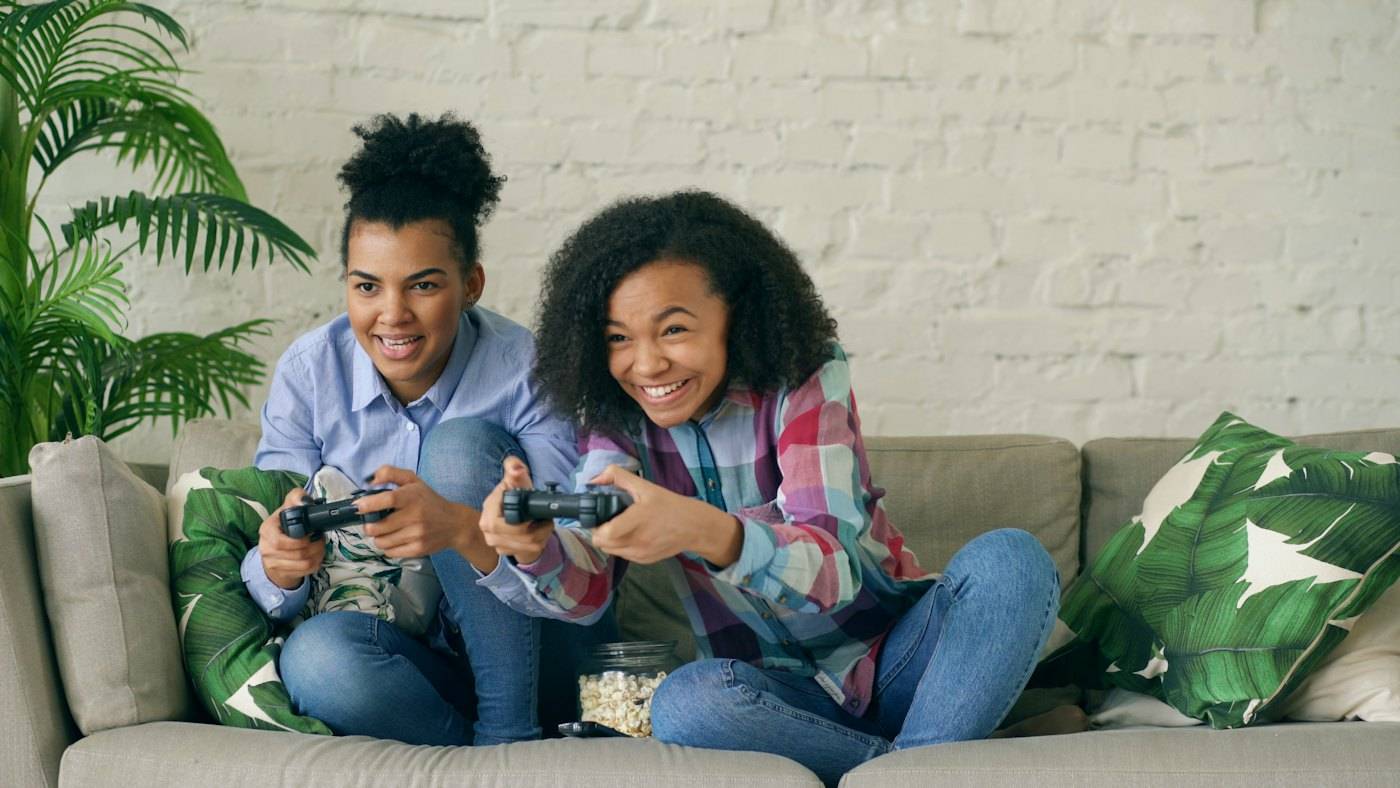 Two girls on a sofa smiling as they play with controllers
